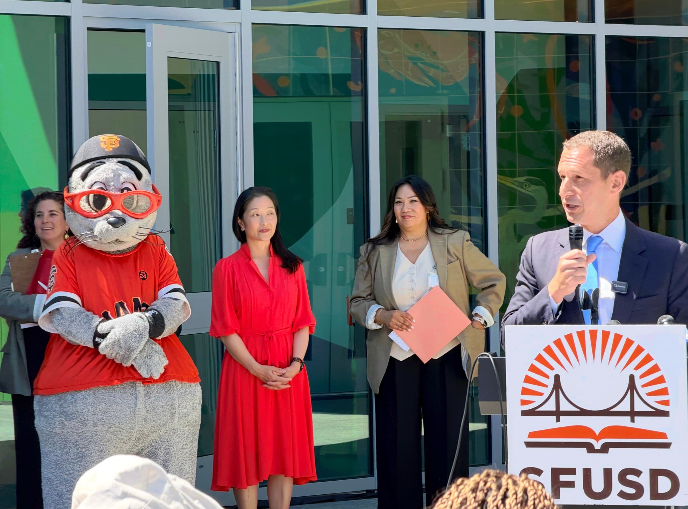Speakers standing beside an SFUSD podium at the ribbon-cutting ceremony, with the Giants mascot alongside them